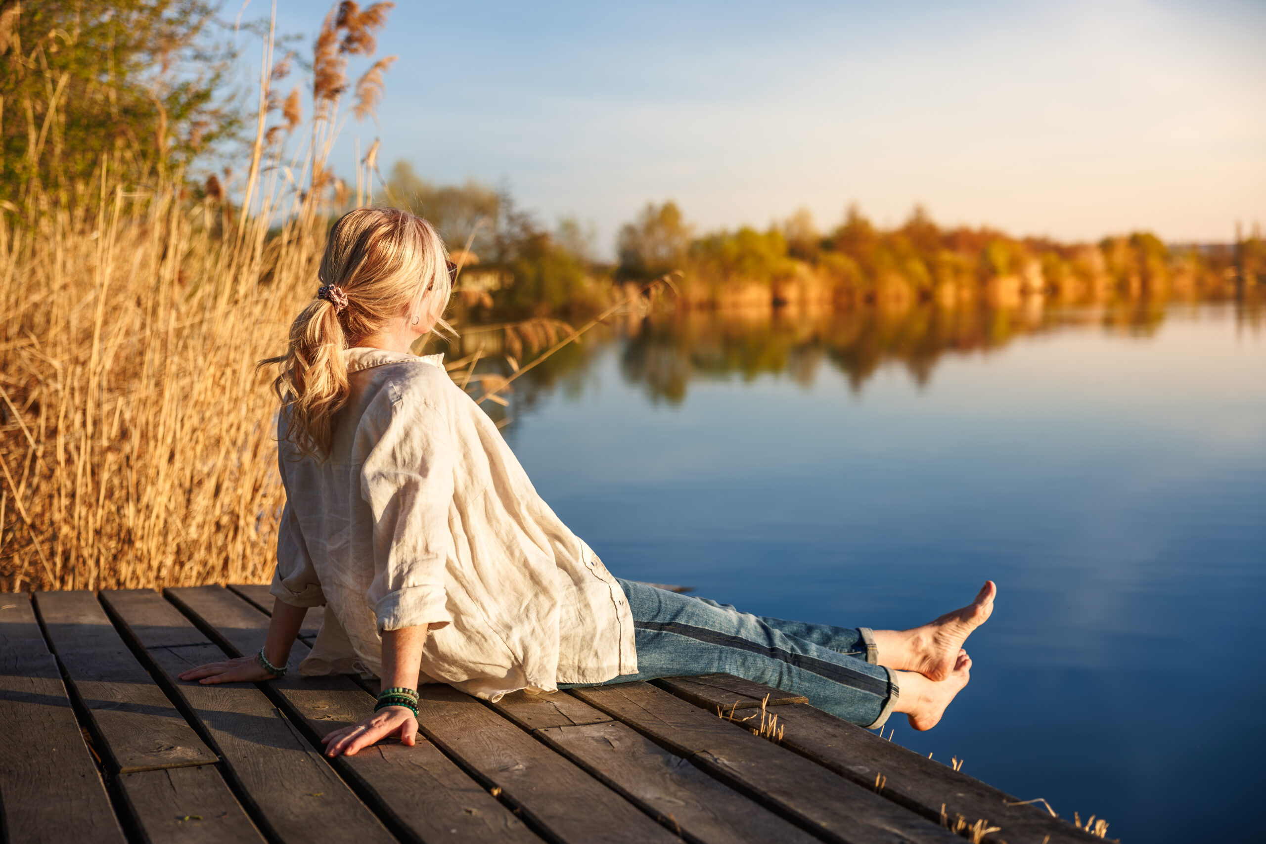Woman sitting and relaxing barefoot on wooden pier by still lake during golden hour of indian summer