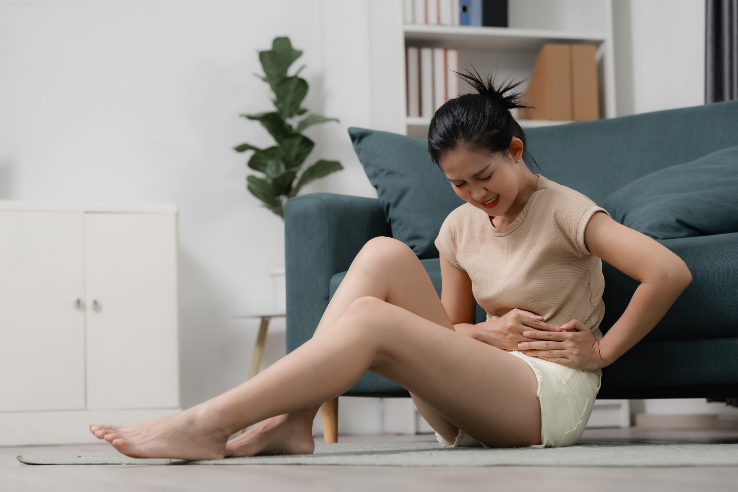 Menstrual cramps and period pain concept showing a woman holding her lower abdomen while sitting on the floor at home in discomfort. period, pain, cramps, menstruation, healthcare